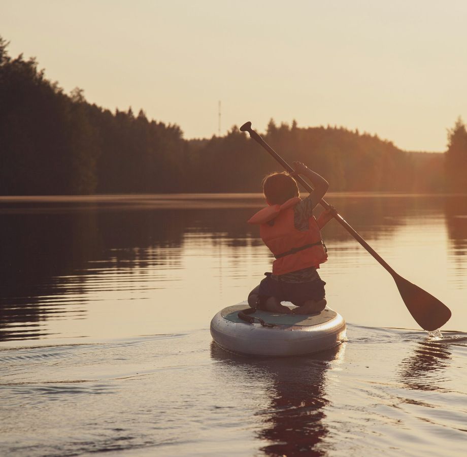 Person paddle boarding on a calm lake at sunset with forested shoreline and warm golden light.